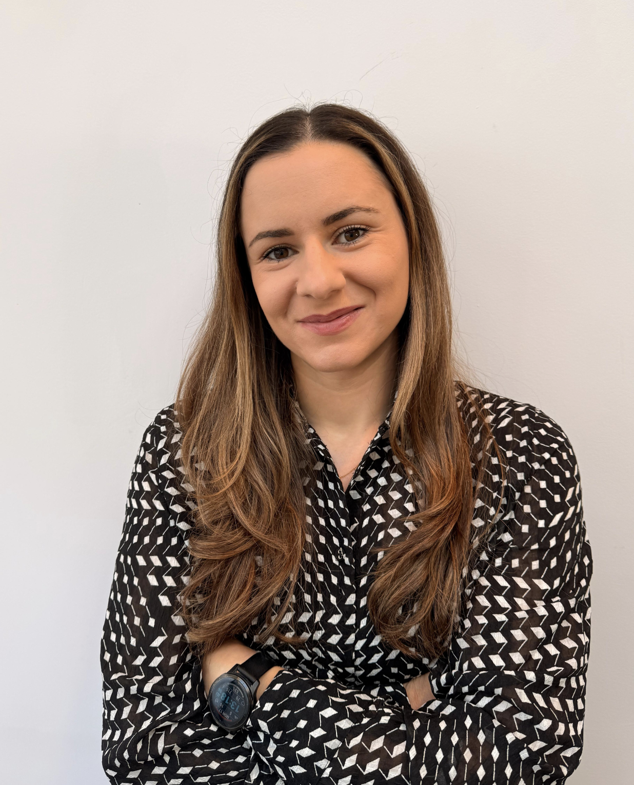 Smiling woman with long brown hair wearing a black and white patterned blouse and a black smartwatch, standing against a plain light background with arms crossed.