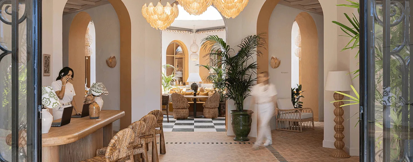 Elegante recepción de hotel con un mostrador de madera, una mujer hablando por teléfono, sillas de mimbre, plantas verdes y mucha luz natural.