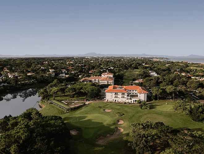 Campo de golf verde con un lago a la izquierda y edificios con techos rojos rodeados de árboles bajo un cielo azul claro.