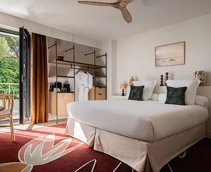 Chambre lumineuse avec grand lit double, coussins verts, porte fenêtre donnant sur un balcon, et mobilier en bois clair.