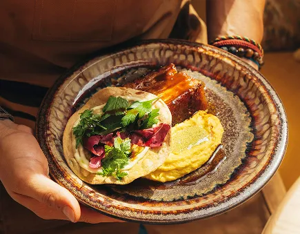 Assiette contenant un morceau de viande braisée, une purée jaune et un wrap garni de feuilles de salade et herbes fraîches, tenue par deux mains.
