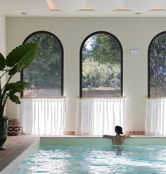 Personne se tenant au bord d'une piscine intérieure avec trois grandes fenêtres arquées donnant sur un jardin verdoyant.