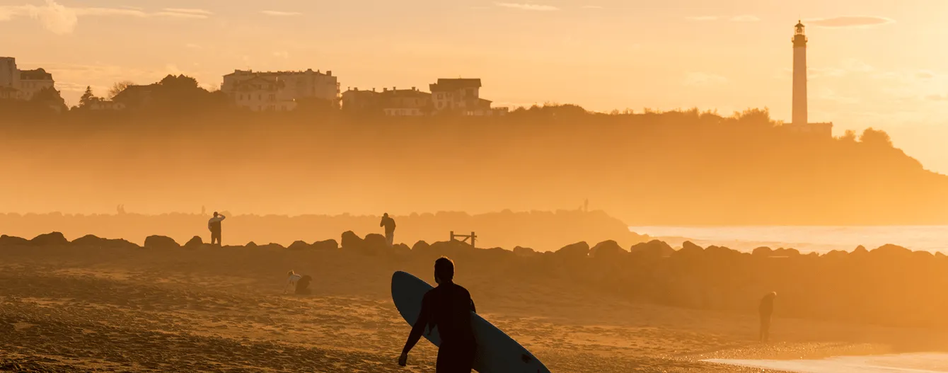 Un surfeur portant une planche de surf marche sur la plage au coucher du soleil avec des silhouettes de personnes et un phare en arrière-plan.