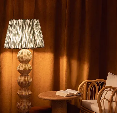 Lámpara de ratán de diseño con pantalla estampada, mesa redonda de madera con libro abierto y sillón de ratán con cojín blanco frente a una cortina marrón.