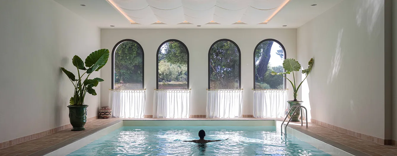 Personne nageant dans une piscine intérieure devant quatre grandes fenêtres en arc donnant sur un jardin verdoyant.