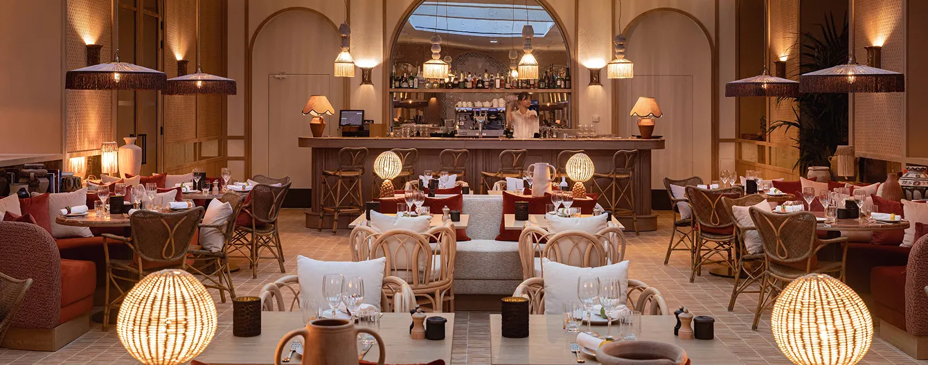 Warm interior of a restaurant with set tables, rattan chairs, decorative lamps and a bar in the background.