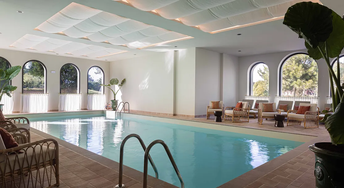 Piscine intérieure rectangulaire avec chaises en rotin et grandes fenêtres à arches laissant entrer la lumière naturelle.