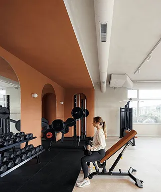 Mujer sentada en un banco de pesas en un gimnasio moderno con mancuernas y grandes ventanales.