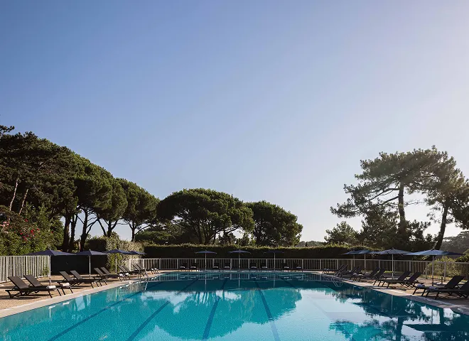 Piscina exterior rodeada de tumbonas y sombrillas con árboles de fondo bajo un cielo despejado.