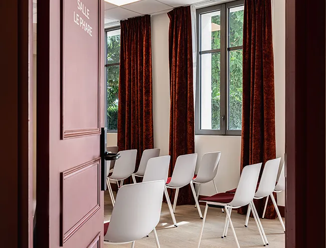 Meeting room with white chairs and red cushions, dark red curtains, and windows that let in natural light.