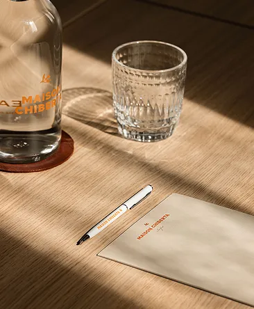 Glass bottle and glass with the inscription Maison Chiberta, pen and paper on a wooden table illuminated by natural light.