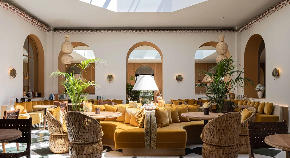 Luminosa sala de estar con sofás curvos amarillos, mesas redondas de madera, sillones de mimbre y grandes plantas verdes.