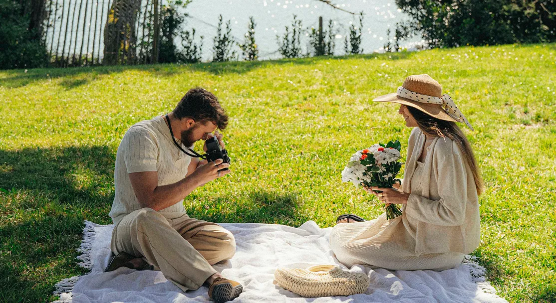 Un hombre toma una foto con una cámara mientras una mujer con sombrero sostiene un ramo de flores sentada sobre una manta blanca en un parque soleado.