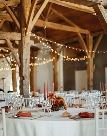 Sala de recepción rústica con vigas de madera, mesas redondas con manteles blancos, decoraciones florales naranjas y velas rosas, vasos rojos y servilletas.