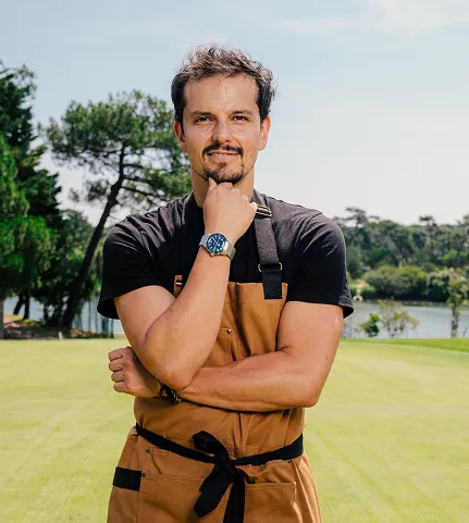 Homme avec une barbe légère portant un tablier marron et une montre bleue, posant dans un parc verdoyant avec des arbres et un lac en arrière-plan.