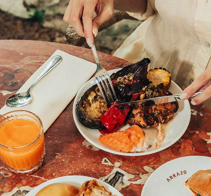 Plate of grilled vegetables and salmon with a glass of orange juice on a stone table, one person holding a fork and a knife.