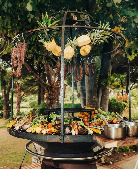 Outdoor grill with hanging pieces of meat and pineapple, surrounded by assorted grilled vegetables on the grill.
