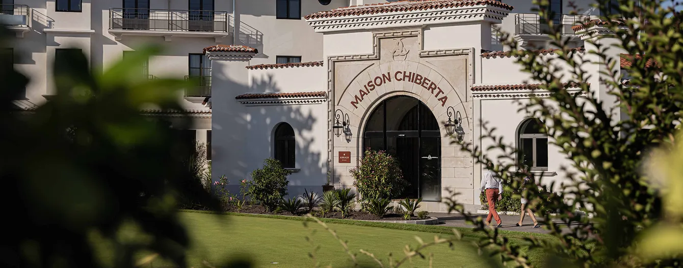 Main facade of the Chiberta House with a stone entrance arch and two people walking in front, partial view through leaves.