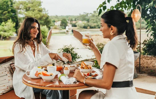 Dos mujeres desayunando al aire libre con bebidas y bollería en una mesa redonda.