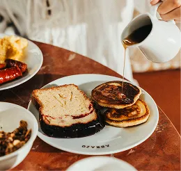 Persona que vierte almíbar sobre una pila de tortitas servidas con una rebanada de pastel y guarniciones sobre una mesa.