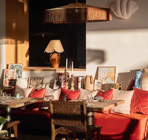 Luminosa sala de estar con sofá y cojines rojos, mesa y silla de madera, decoración cálida con lámparas y cuadros.