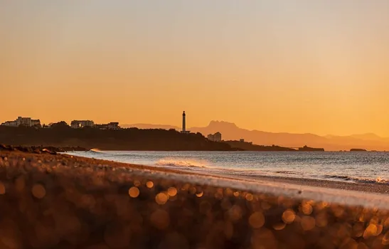 Playa con un faro en la distancia al atardecer, cielo anaranjado y montañas al fondo.