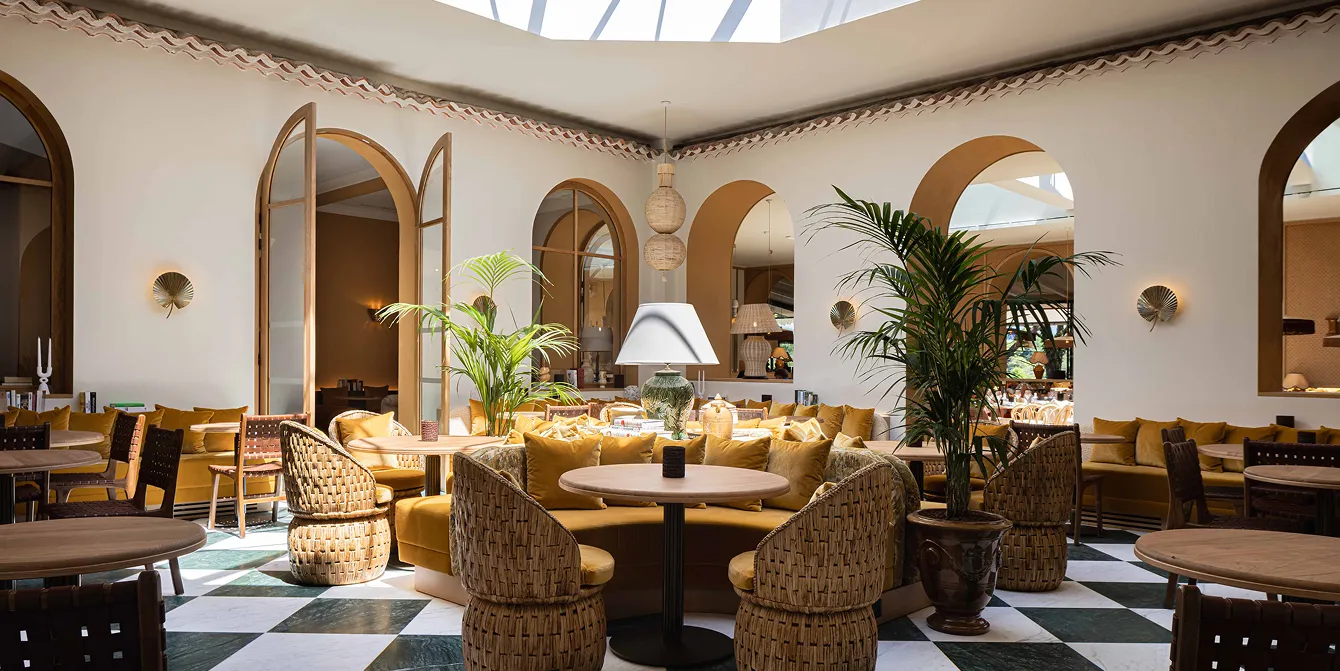 Bright cafe interior with rattan seats, yellow cushions, large green plants, and black and white checkerboard flooring.