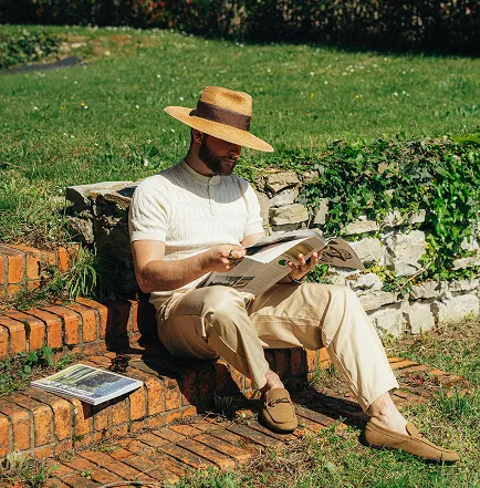 A man wearing a straw hat and a white sweater is sitting on brick steps outside, reading a book, with another book lying next to him.