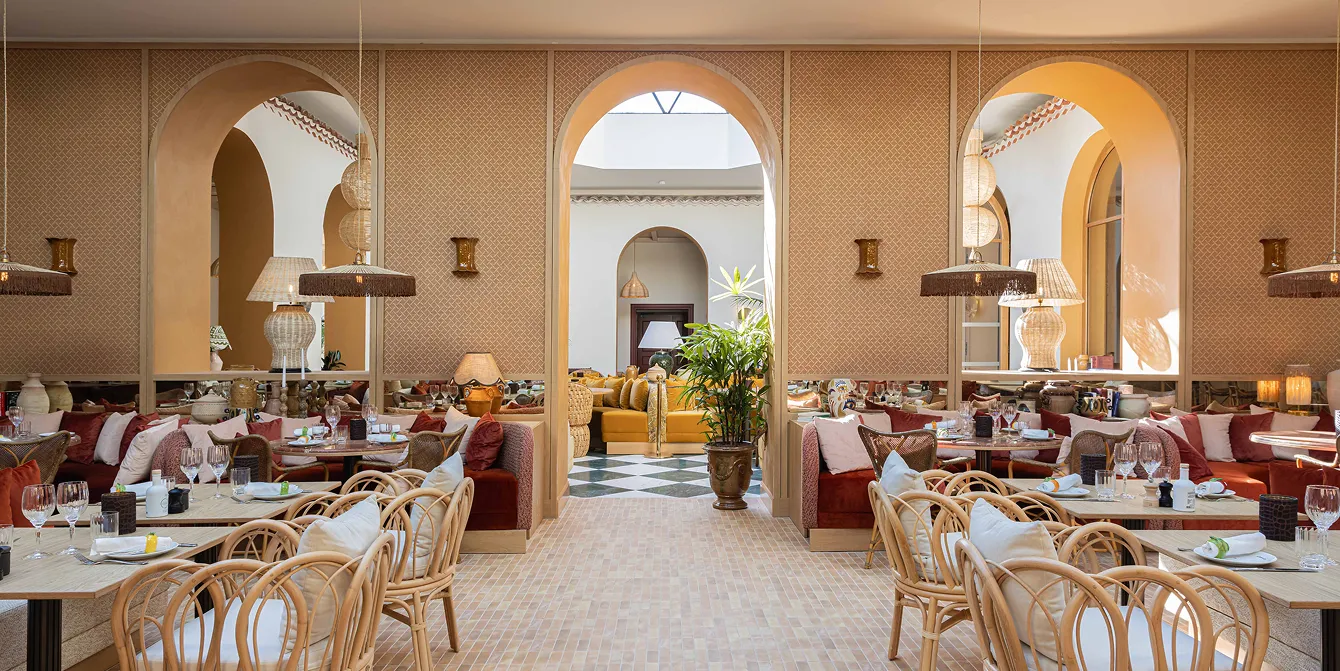 Intérieur élégant d'un restaurant avec arches beige, tables en bois avec verres et serviettes, chaises en rotin et canapés rouges avec coussins, décorations et plantes.