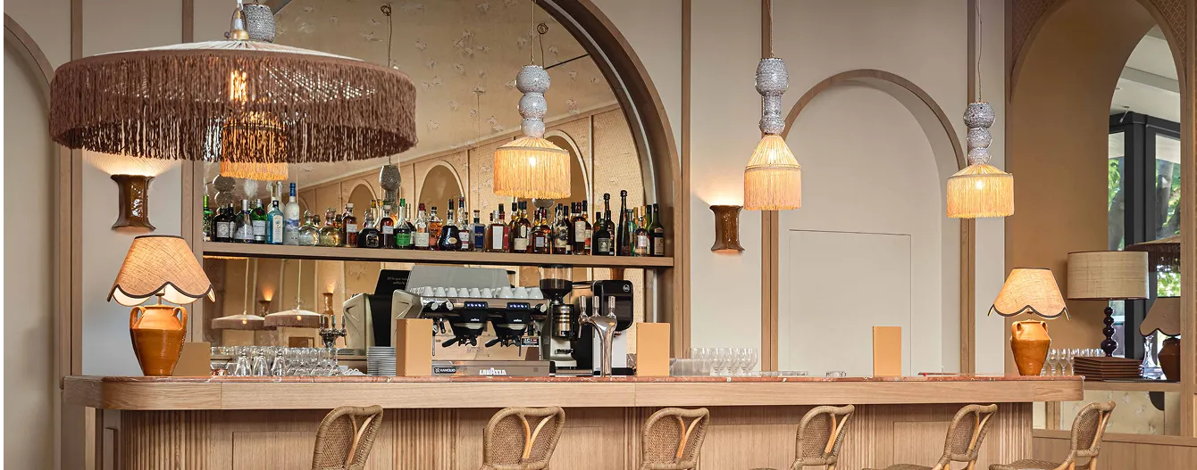 Bar en bois clair avec tabourets en rotin, éclairage suspendu à franges et étagères de bouteilles derrière un grand miroir arcade.