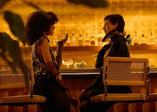 Two elegant women chat sitting on stools at the bar in a warm and subdued atmosphere.
