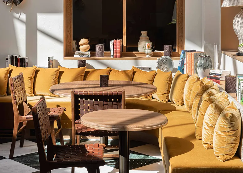 Bright sitting area with a mustard-yellow corner sofa lined with cushions and two round wooden tables surrounded by chairs with woven seats.