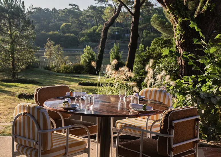 Round wooden table with four striped chairs and glasses set outside in a sunny garden with trees in the background.