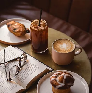 Table with an open book and glasses, iced coffee, latte with heart latte art, chocolate croissant, and muffin with icing.
