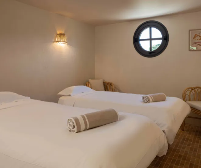 Chambre avec deux lits simples blancs, serviettes roulées à rayures sur chaque lit, fenêtre ronde, chaise en rotin et lampe murale.