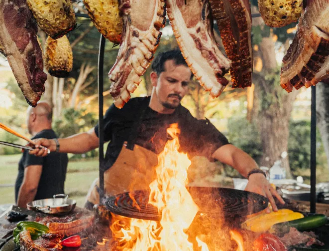 Chef in apron cooking suspended meat and vegetables on an outdoor high heat barbecue.