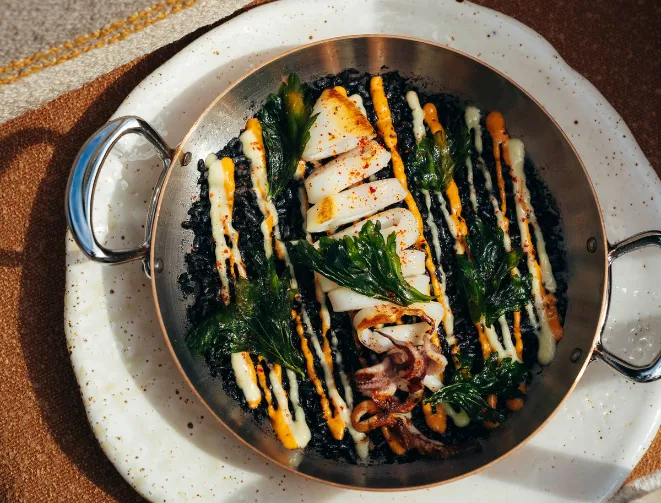 Grilled calamari dish served with green leaves and orange and white sauces, served in a round pan with handles.