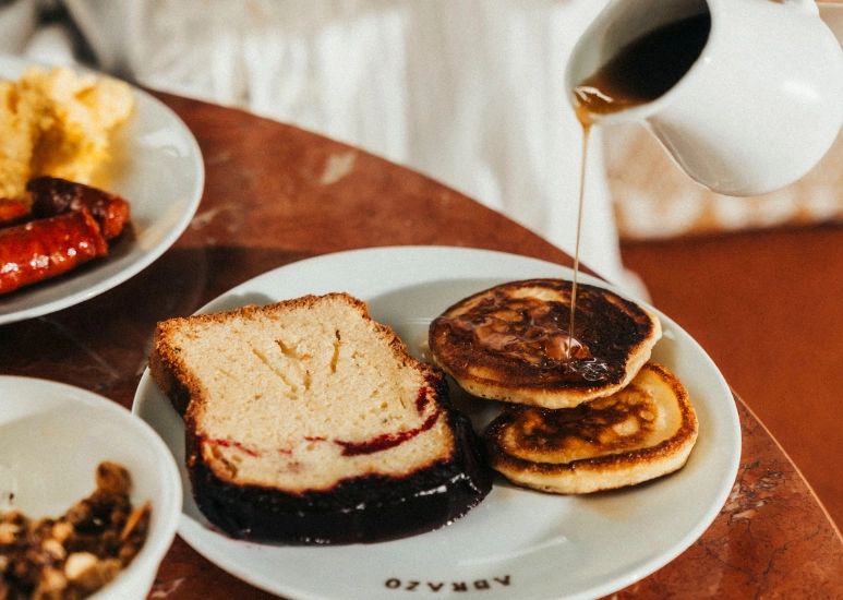 Sirop versé sur deux pancakes avec une tranche de gâteau sur une assiette blanche sur une table en bois.