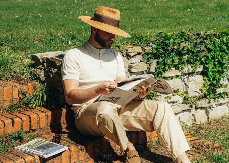Homme assis sur des marches en briques, portant un chapeau de paille et lisant un magazine par une journée ensoleillée.
