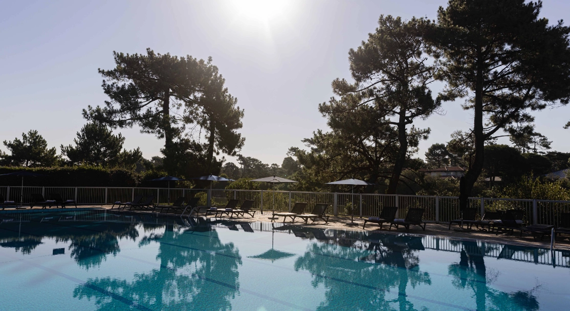 Outdoor pool surrounded by sun loungers and umbrellas with trees in the background at sunset.