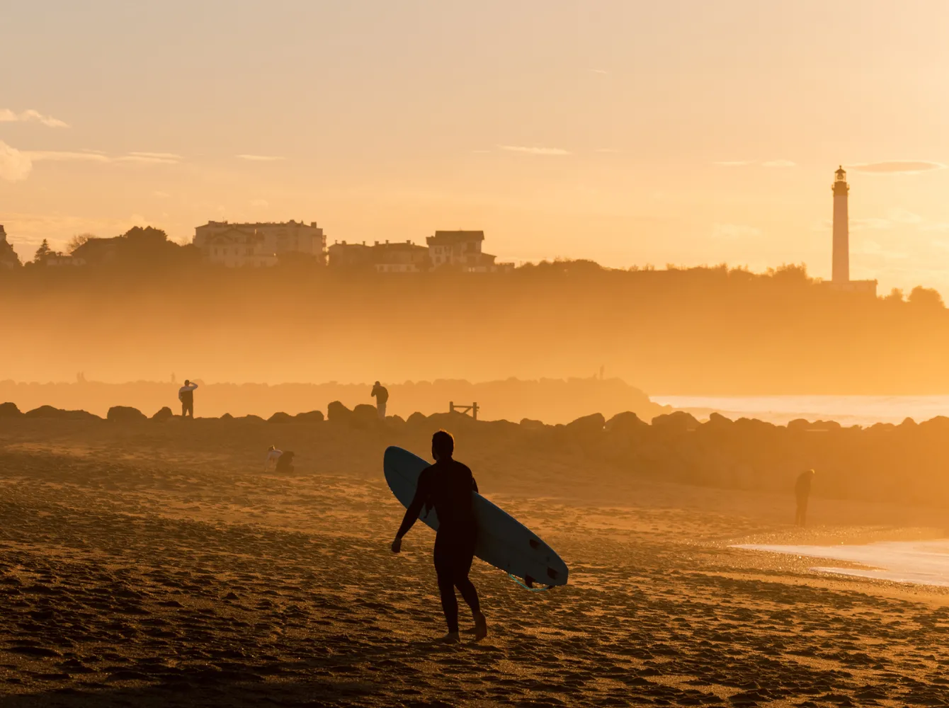 Surfeur avec planche marchant sur la plage au coucher du soleil à Anglet, phare en arrière-plan