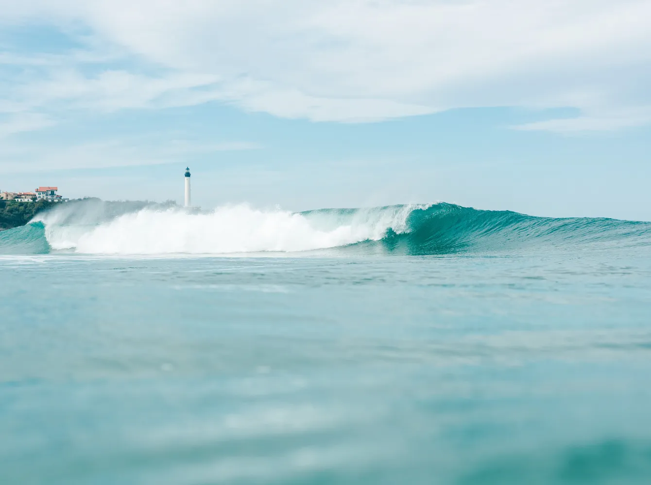 Vague déferlante turquoise sur la côte du Pays basque avec phare et maisons au loin