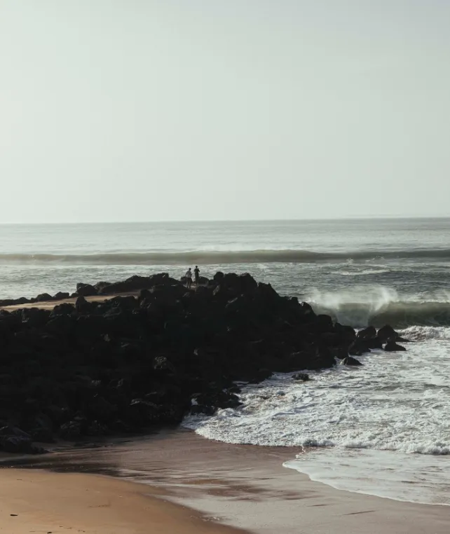 Deux personnes sur une jetée rocheuse face à l’océan à Anglet, vagues sur les rochers et plage au premier plan