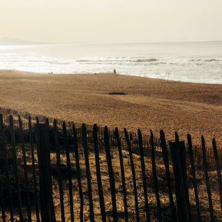 Plage avec clôture en bois au premier plan et vagues douces sous un ciel clair au loin.