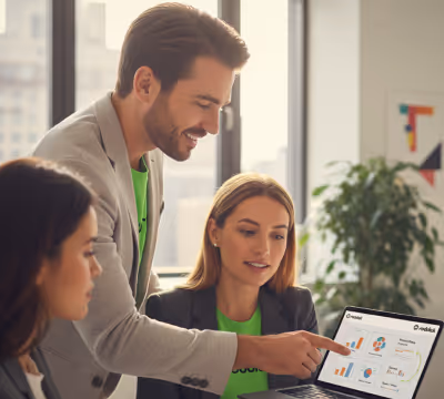 A man points at a laptop screen displaying charts while two women watch attentively in a bright office.