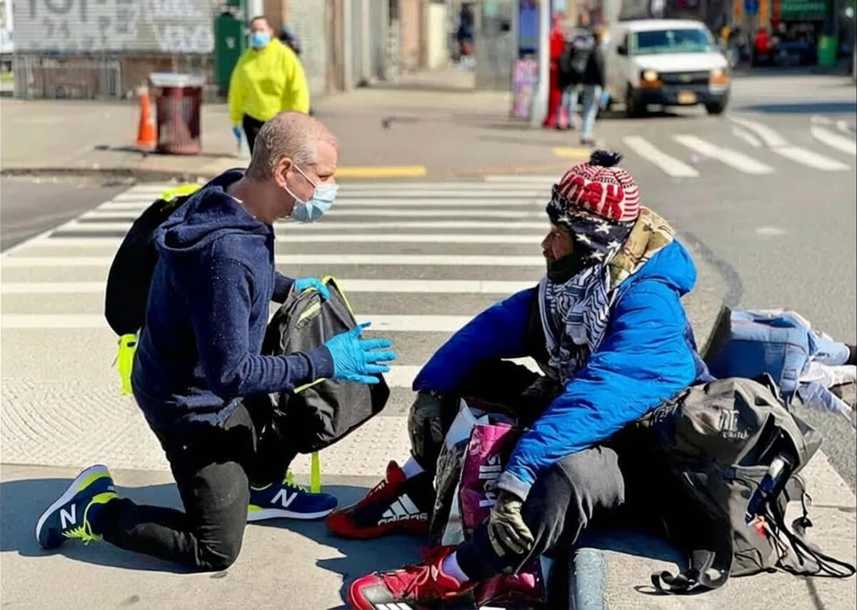 A masked volunteer kneels on a city sidewalk, talking with a bundled-up person seated at a street corner, suggesting an outreach or assistance moment.