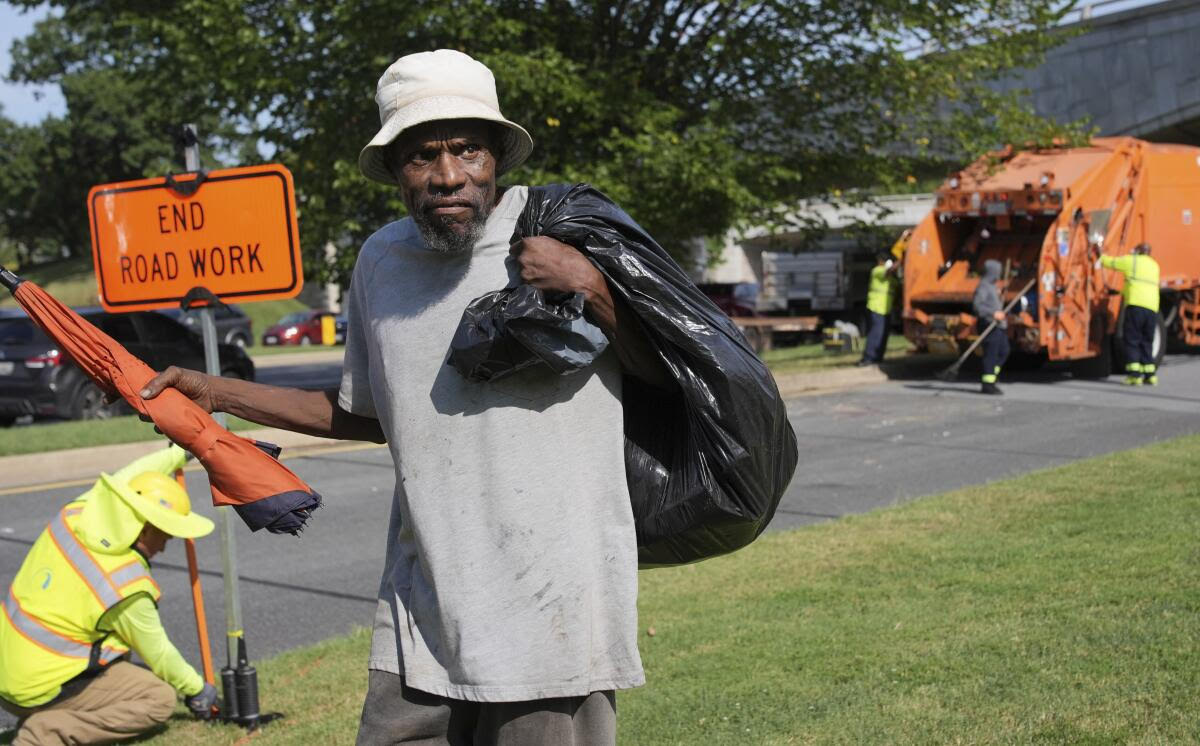 A man stands near a roadside holding a folded umbrella and a black bag, with road work signs and sanitation workers in the background.