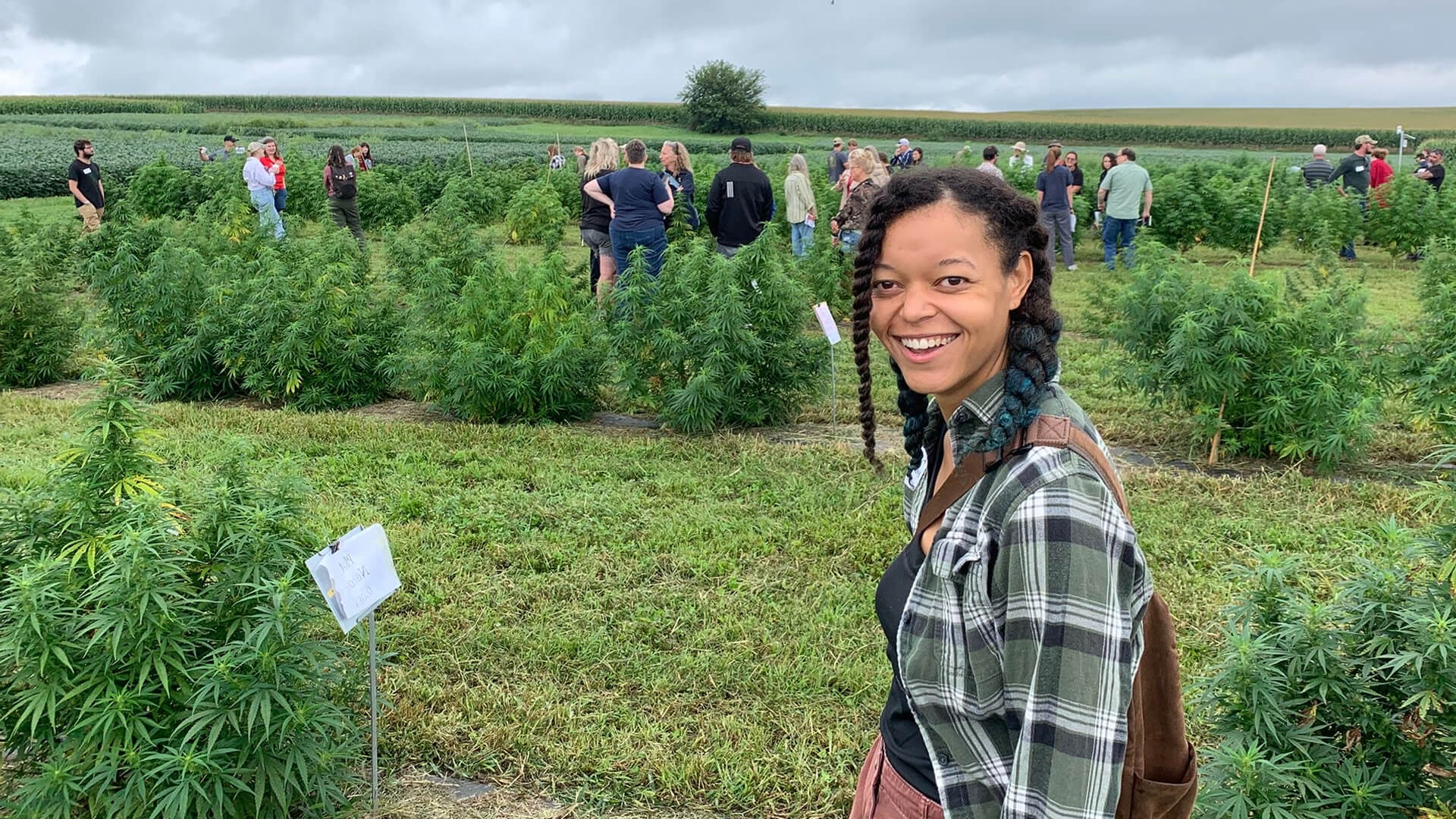 Smiling person standing in a hemp field with rows of plants and people in the background.