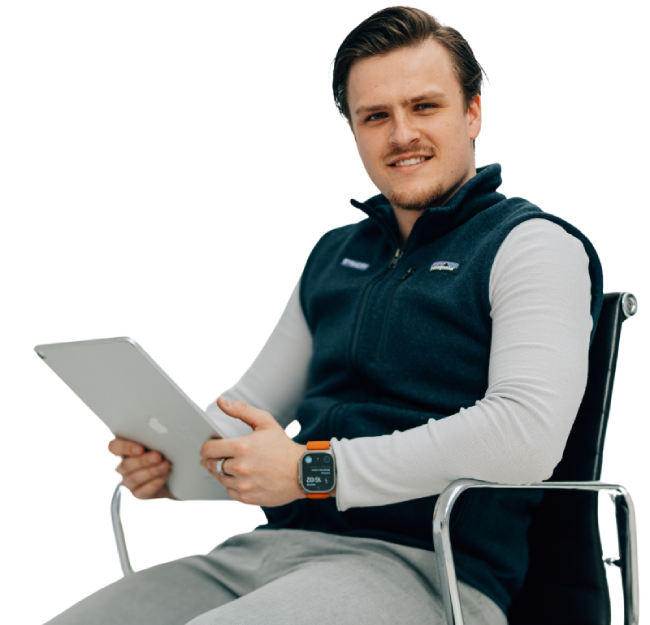 Ein junger Mann sitzt auf einem Stuhl und hält ein Tablet, trägt eine dunkelblaue Weste und eine Smartwatch mit orangem Armband.