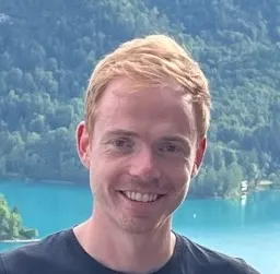 Junger Mann mit Bart und kurzen Haaren lächelt vor grauem Hintergrund.